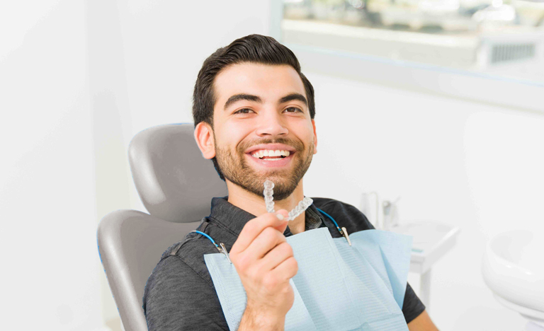 Smiling Man Holding Clear Aligner Sitting In Orthodontist Chair Scaled