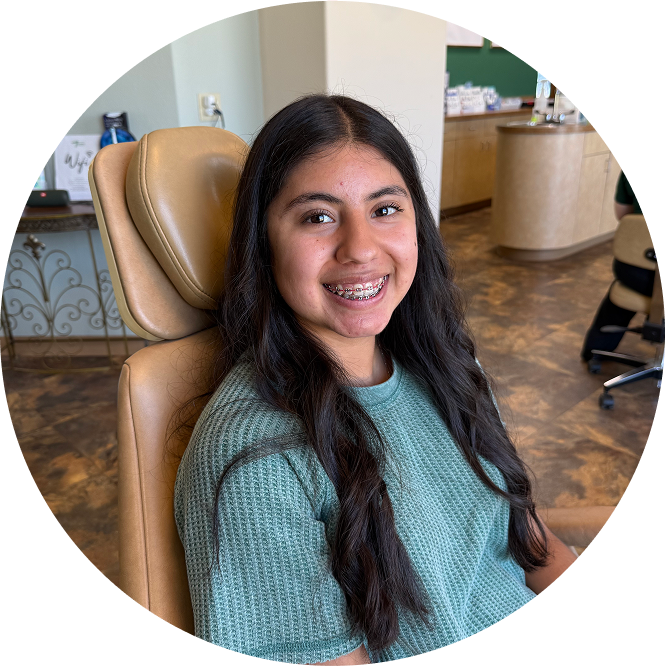 A girl with brown hair, smiling with braces sitting in an examining chair at the Tennison Orthodontics office.