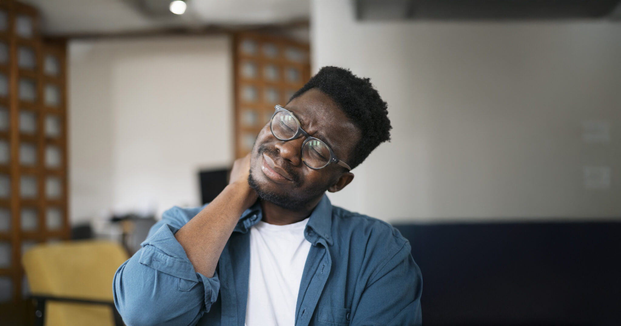 Young man sitting at home experiencing and dealing with the pain in his neck, expressing his discomfort while holding his hand on a painful place